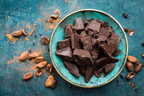 Dark chocolate pieces arranged on a white plate, with cocoa beans scattered around.